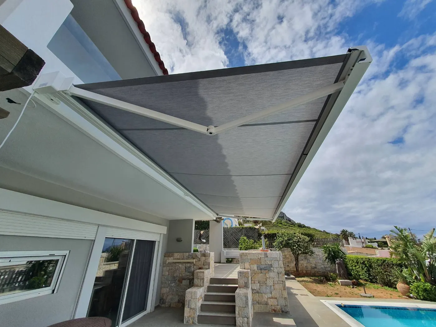 Retractable grey fabric awning extended over a villa terrace with garden and pool behind
