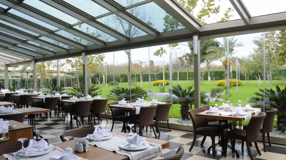 Restaurant terrace under a glass roof with garden views through floor-to-ceiling glass walls