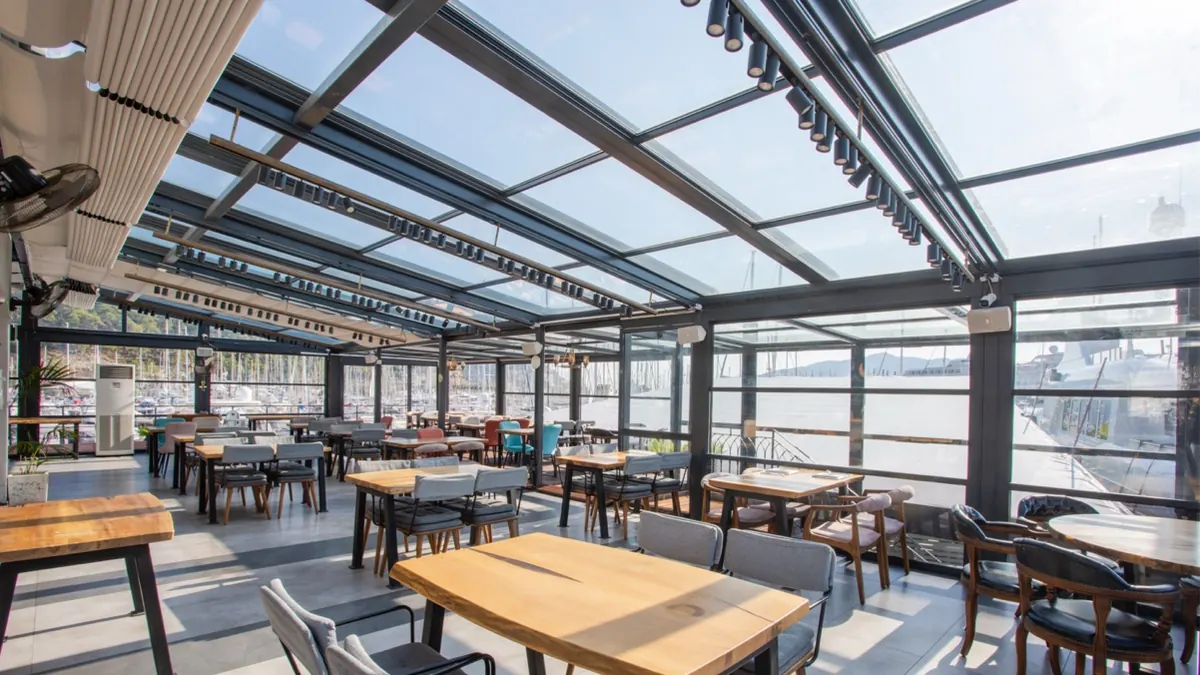 Marina restaurant dining area under an angled glass roof with spotlights and harbour views