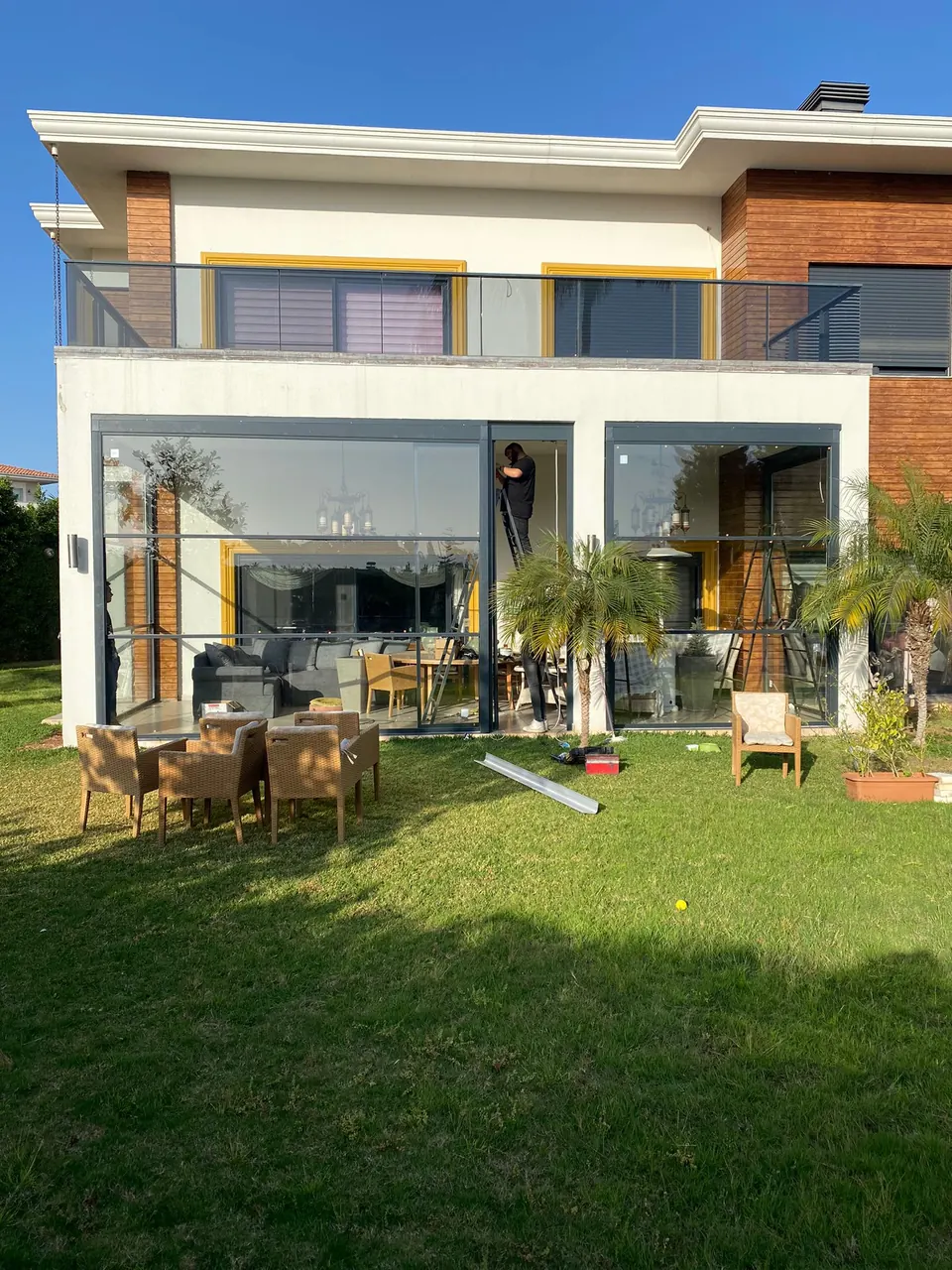Garden-side villa terrace enclosed by dark-framed guillotine glass panels while an installer works on a ladder
