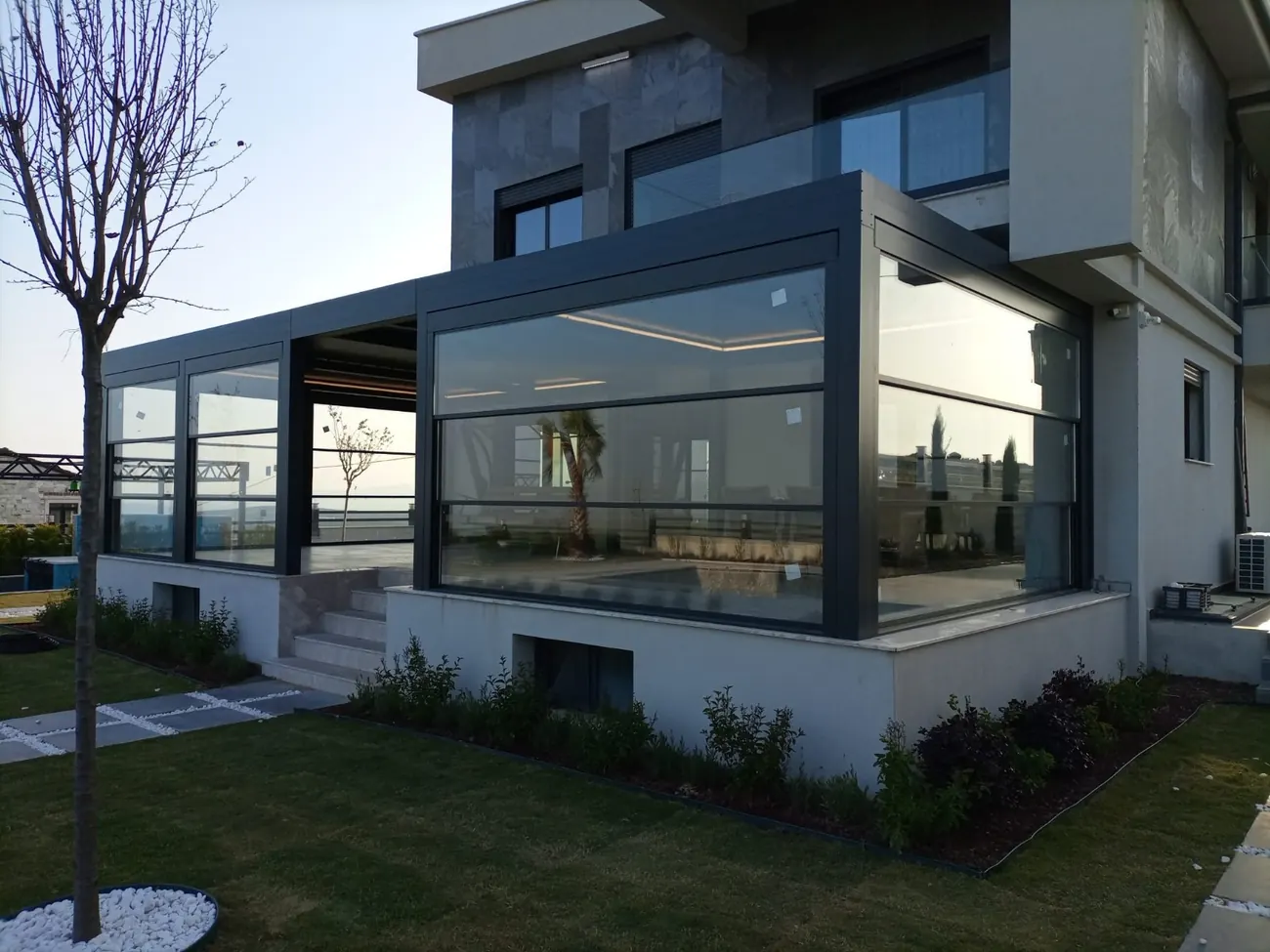 Café terrace enclosed by a dark-framed guillotine glass system with lawn and sea horizon behind