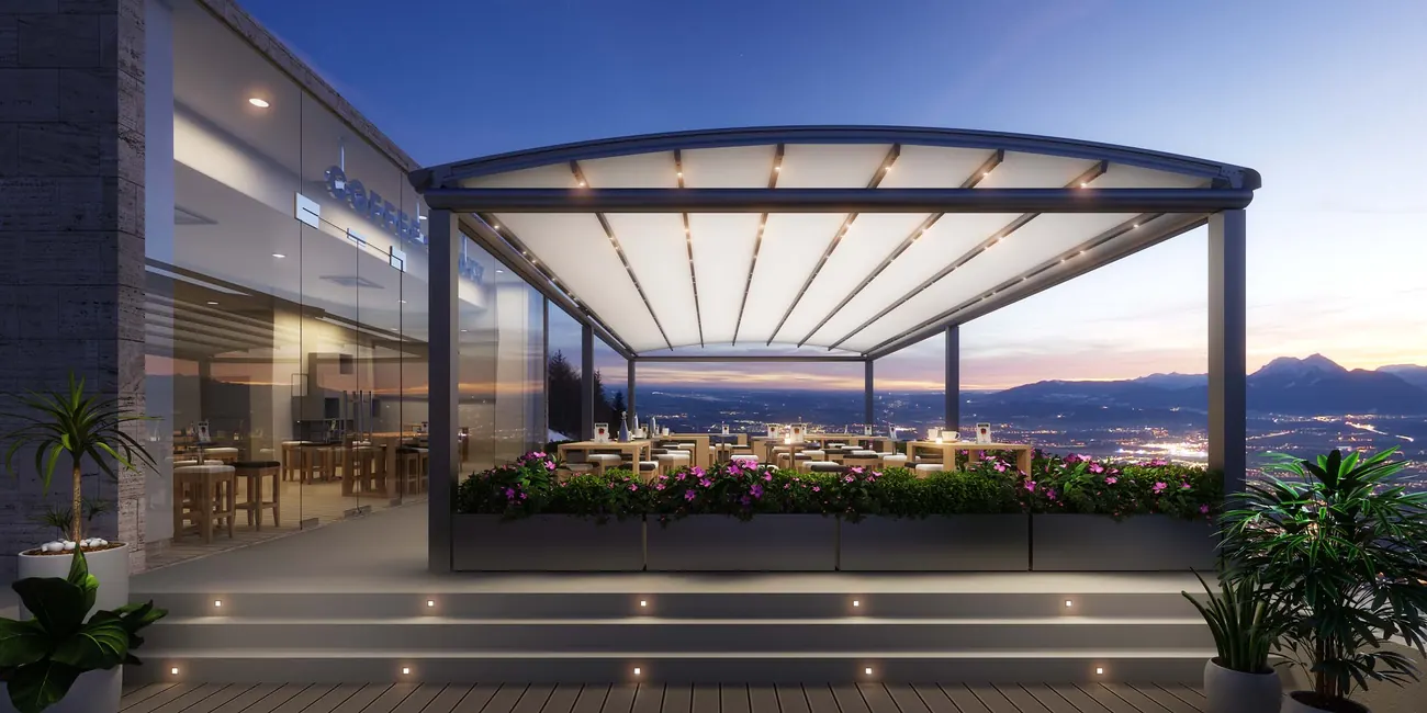 Arched canopy with warm lighting over an outdoor seating area at dusk with glass screens on the sides
