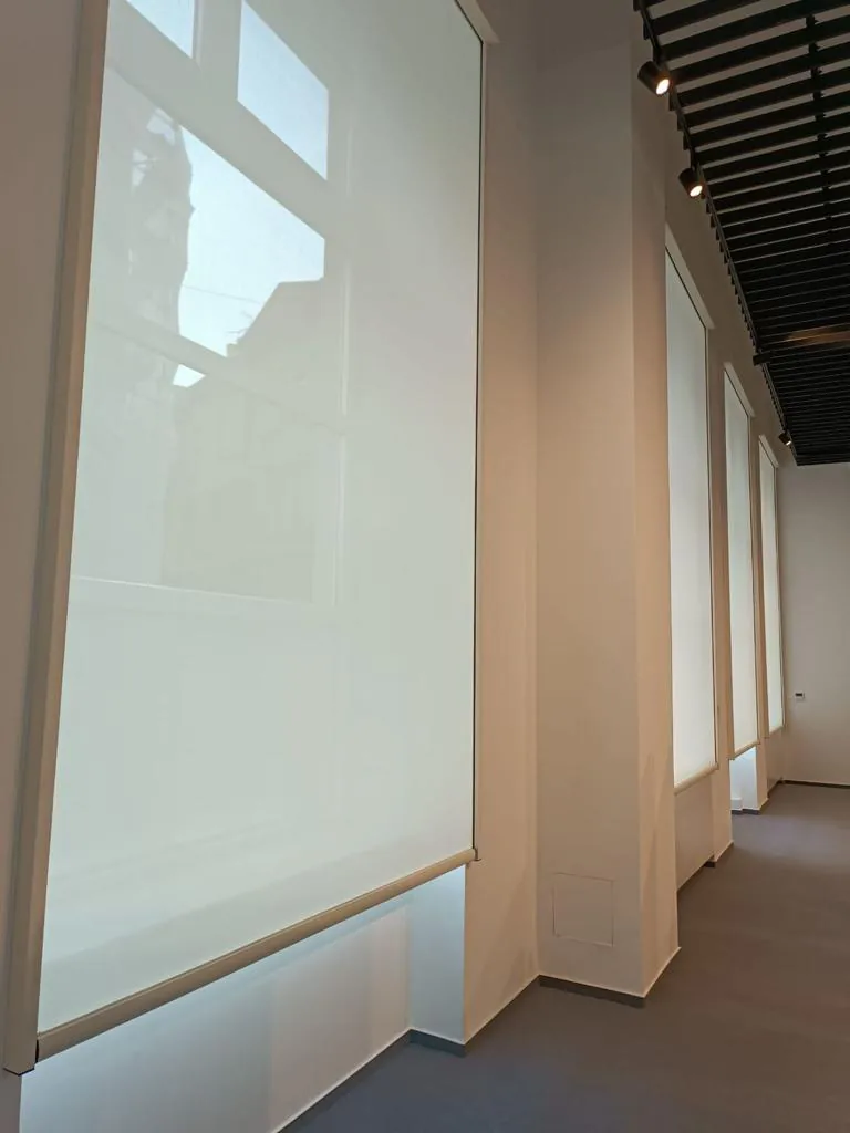 Full-height light roller screen over a tall window in a bright hall with dark ceiling beams