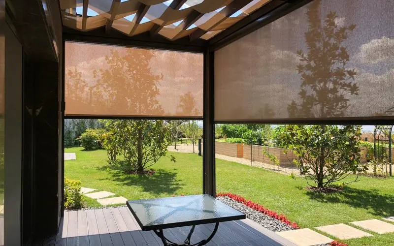 Lowered mesh zip screens on a pergola terrace, filtering sun above a garden lawn